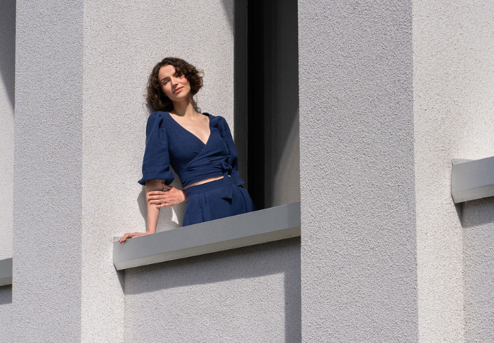 A woman wearing a dark blue two-piece linen outfit from Menique stands on a balcony between light gray modern architectural walls, looking toward the sun.