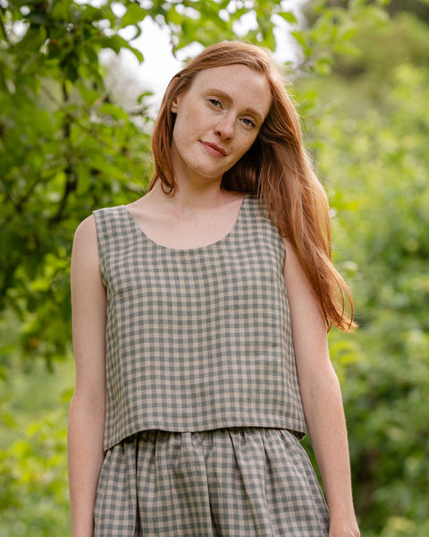 The model in the photograph is visible from the front, with a close-up view of the ANA cropped linen blouse in a green gingham pattern. The top is sleeveless, matched with same garment bottoms. There are blurred nature elements visible in the background.
