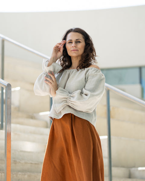 A woman with brown hair, wearing a light beige off-the-shoulder top with puff sleeves and a long rust-brown skirt, poses on concrete stairs.