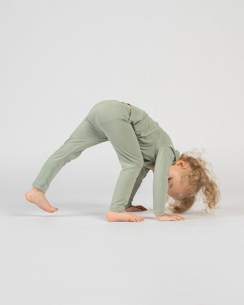 Child in bamboo green long-sleeve outfit playfully bending forward with hands and feet on the floor, forming an inverted V-shape, with curly hair hanging down against a plain background.