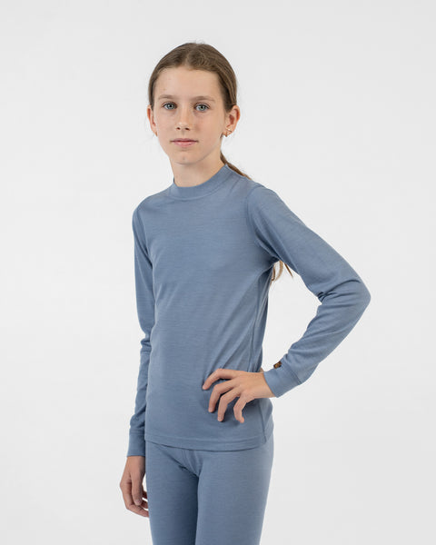 A young girl seen from the thighs up, wearing a dusty blue merino long-sleeve top and matching bottoms, standing with her left hand on her hip.
