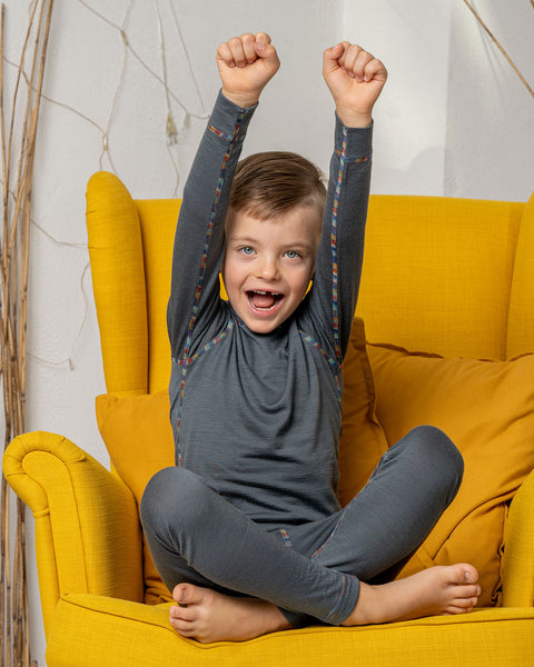 A boy sitting in a yellow armchair, wearing the menique Kids' 160 Merino Pants in Perfect Grey color. He has his hands raised above his head, being cheerful.