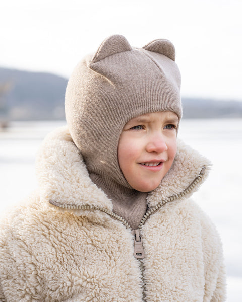 A close-up shot of a young child wearing a creamy beige menique knit balaclava with two ear-like protrusions on top. They are also wearing a cream-colored, fuzzy or shearling-like zippered jacket. The child is looking slightly to their right with a neutral expression. The background is blurred, showing a light, possibly snowy or icy, outdoor environment.
