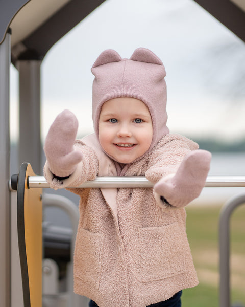 A young child wearing a menique dusty pink balaclava with ears, matching mittens, and a fuzzy pink coat smiles at the camera. They are standing at a metal bar on a playground structure. In the background, there is a blurred body of water and some greenery.