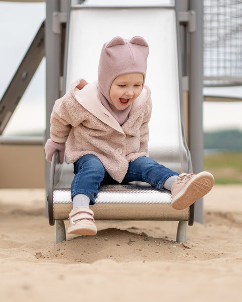 A young child, dressed in a pink textured jacket with a hood, a dusty pink menique balaclava with ear-like protrusions, pink mittens, blue jeans, and pink sneakers, is at the bottom of a silver playground slide. The child is smiling widely with an open mouth, looking down and to their right, seemingly delighted after coming down the slide. Their feet are extended in front of them on the sandy ground, and the metal structure of the slide is visible behind them.