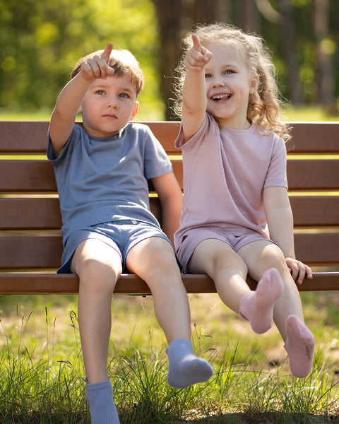 Two children, a boy and a girl, sitting side-by-side on a wooden park bench. The boy is dressed in a blue top and shorts with matching light blue ankle socks, while the girl is wearing a pink top and shorts with matching dusty pink ankle socks. Both are smiling and pointing excitedly up at something out of frame.