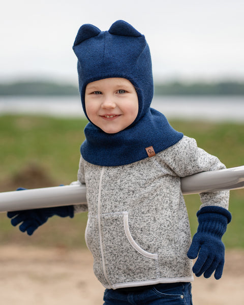 A young child with fair skin and light-colored hair is smiling at the camera, leaning on a light-colored horizontal bar outdoors. They are wearing a grey speckled zip-up jacket, blue jeans, and dark blue knitted menique accessories, including a balaclava-style hat with two rounded ears on top and a pair of ribbed gloves. In the soft-focus background, there's a body of water and a faint green shoreline.