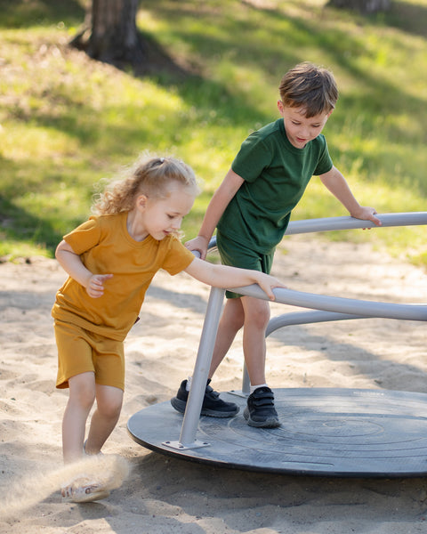 Two children outdoors wearing matching Merino wool T-shirts and shorts in yellow and dark green. The lightweight, breathable kids' outfits are showcased in a sunny park setting, perfect for everyday play, family outings, and warm-weather adventures.