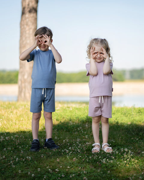 Two young children, a boy and a girl, stand side-by-side in a park setting, both holding their hands up to their eyes to form circle "glasses." The boy is on the left, wearing a blue t-shirt and shorts set with black sneakers. The girl is on the right, wearing a light pink t-shirt and shorts set with white sandals.