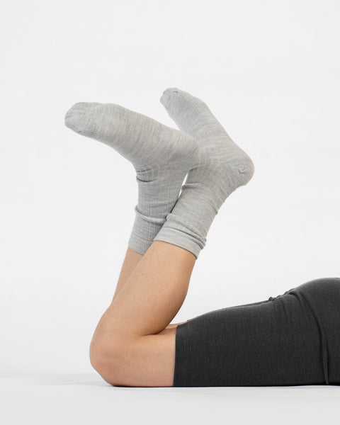 A person lying on their stomach with lower legs raised, wearing light gray ribbed merino crew socks, shown from the side.