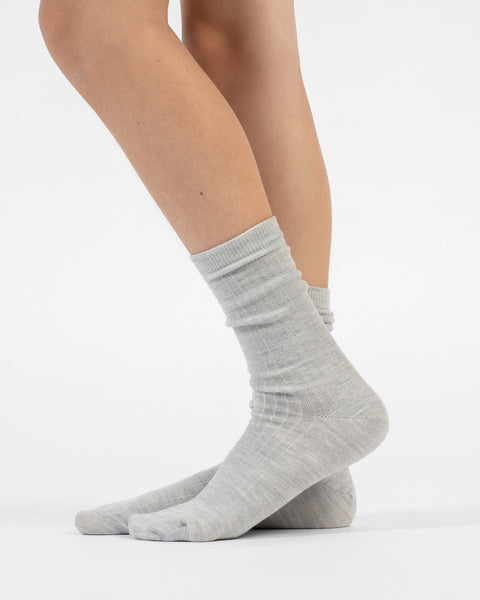 A side-profile view of a person standing, wearing light gray ribbed merino crew socks, with the back foot slightly lifted.