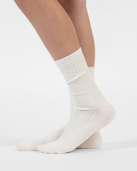 A side-profile view of a person standing, wearing natural ribbed merino crew socks, with the back foot slightly lifted.