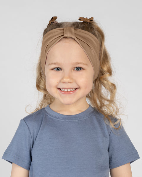 A close-up studio portrait of a young blonde girl with a wide smile looking directly at the camera against a white background. She is wearing a dusty blue short-sleeved t-shirt and a beige ribbon headband, with small brown hair bows visible in her hair behind the headband.