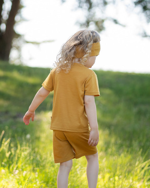 Young girl walking through a grassy meadow wearing a yellow Merino wool T-shirt, matching shorts, and a coordinating Merino wool headband. The soft, breathable outfit is showcased in a sunny outdoor setting, offering comfortable everyday wear for active kids.