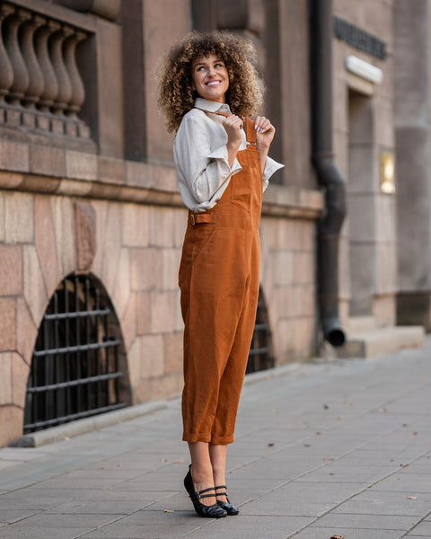 Woman with curly hair standing in a street and wearing Linen Pinafore Jumpsuit Nicci Almond Brown