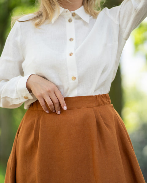 A close-up view highlights the textured fabric and wooden button details of a long-sleeve pure white linen shirt. The shirt is elegantly tucked into the elastic waistband of an almond brown skirt, showcasing a classic and natural styling.