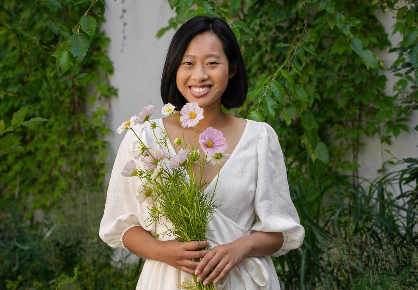 A smiling woman in a white linen dress from menique holds a bouquet of pastel-colored cosmos flowers, standing in front of a green, leafy garden wall.