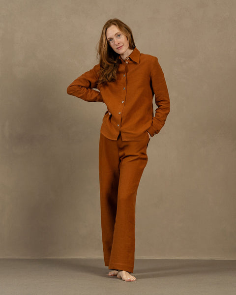 A barefoot woman with long, light brown hair stands on a light brown surface in front of a muted brown wall. She is wearing an almond brown-colored, long-sleeved linen button-down shirt and matching wide-legged linen pants, with one hand in her pocket and the other resting on her hip.