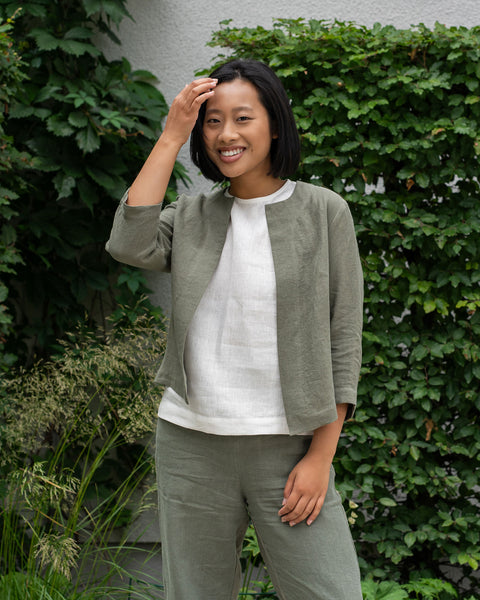A woman wearing a white top, gray jacket, and gray pants poses outdoors against a green leafy background.