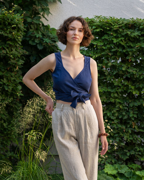 A light-skinned person with short, wavy brown hair is standing outdoors with one hand on their hip and a neutral expression. They are wearing a storm blue sleeveless wrap top tied at the side and natural linen pants. Lush green foliage serves as a backdrop.