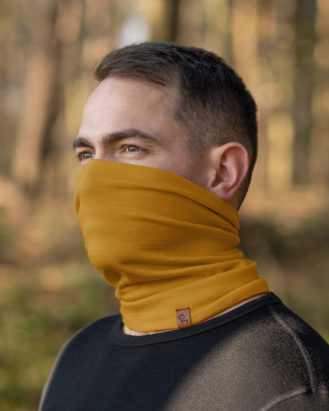 A person with short, dark hair and light eyes is shown wearing a spicy yellow neck gaiter that covers their mouth and nose. They are also wearing a black long-sleeved shirt, and the blurred background suggests an outdoor setting with trees.