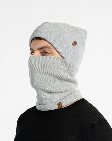 A male model, viewed in profile, wears a light gray beanie and a matching neck gaiter that covers his nose and mouth. Both the beanie and gaiter feature a small, brown leather patch with a Menique logo. He has a short beard and dark hair. The background is solid white.