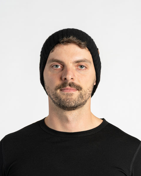 A person with light skin, short brown hair, a mustache, and light-colored eyes is shown in a headshot against a white background. They are wearing a black knit beanie with a small brown tag on the side and a black long-sleeved shirt. They are looking directly at the camera with a neutral expression.