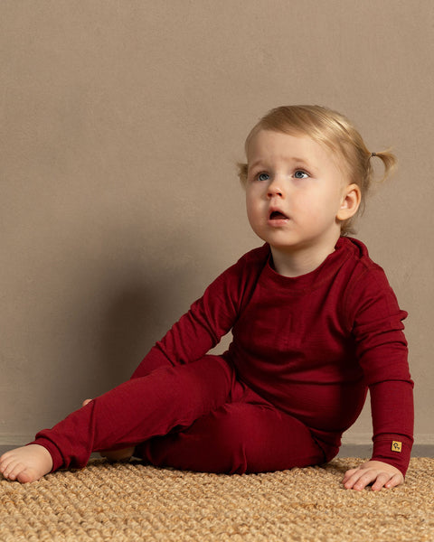 A toddler sitting on the ground, looking up and wearing cheerry red merino wool set.