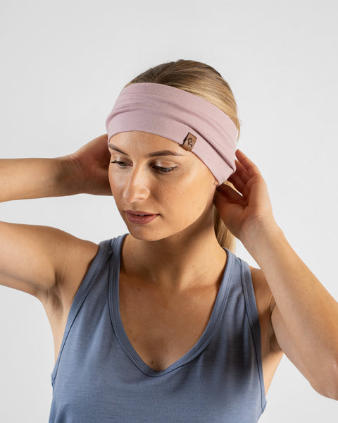 A studio portrait of a blonde woman looking downward and to the left. She is wearing a dusty blue sleeveless top and a dusty pink headband, with both hands raised behind her head to adjust the band against a light grey background.