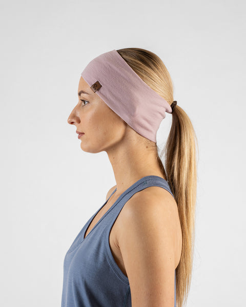 A side profile studio portrait of a blonde woman facing left against a plain light grey background. She is wearing a dusty blue sleeveless top and a wide dusty pink headband over her hair, which is tied back in a long ponytail. A small brown logo tag is visible on the side of the headband.