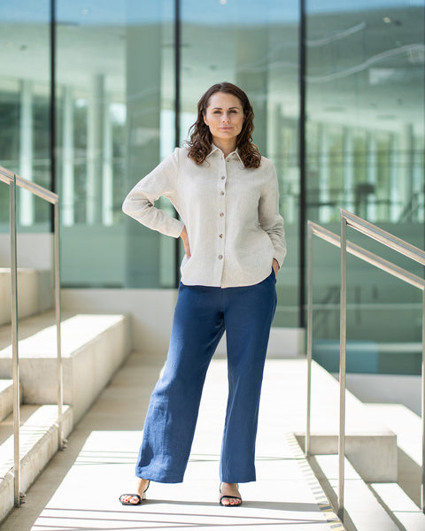 A woman with brown hair, wearing a natural linen button-front shirt and wide-leg white trousers, stands outdoors against a modern concrete structure.