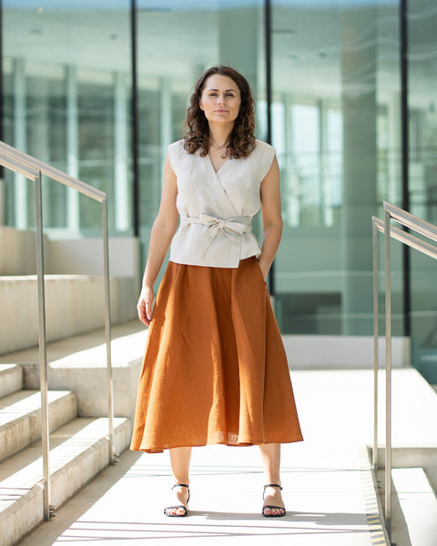 A woman with shoulder-length wavy brown hair stands outdoors on concrete steps, facing forward with her hands in her pockets. She is wearing a natural wrap top with a V-neck and a tied waist, paired with a long, linen skirt that reaches mid-calf. She also wears dark sandals.