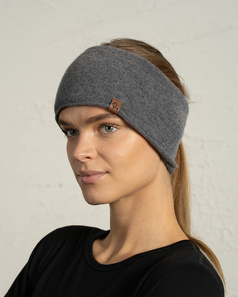 A woman with her hair tied back in a low ponytail wears a wide, soft gray knit headband that covers her ears. She is dressed in a black top and faces slightly to the side, showing the headband’s curved shape and a small brown logo tag. The light, textured wall behind her provides a neutral backdrop.