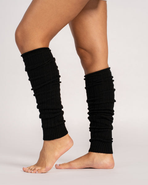 A person stands barefoot, wearing a pair of black, ribbed-knit leg warmers that are scrunched down their calves. The photo is taken from the side against a plain, light-colored background.