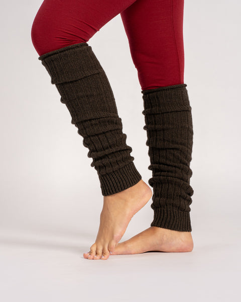 A person stands barefoot, wearing brown, ribbed-knit leg warmers scrunched down over red leggings. The photo is a side view of their lower legs against a solid light gray background.