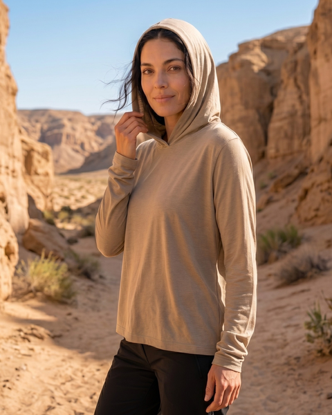 An outdoor portrait of a woman standing on a sandy path within a rocky canyon under a clear blue sky. She is looking directly at the camera with a gentle smile, wearing a lightweight beige long-sleeved hoodie with the hood pulled up and black pants, with one hand resting near the edge of the hood.
