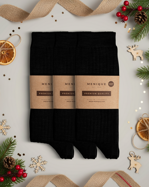 Three pairs of black ribbed socks neatly arranged with kraft paper packaging, surrounded by festive Christmas decor like pine branches, dried oranges, and ornaments.