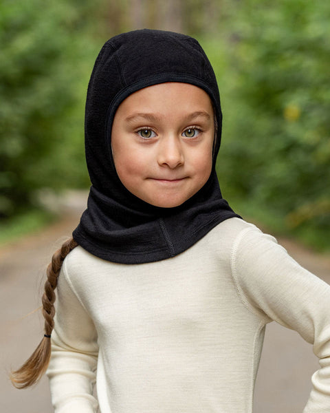 A girl wearing the menique kids' merino 160gsm balaclava in black color, a forest background picture.