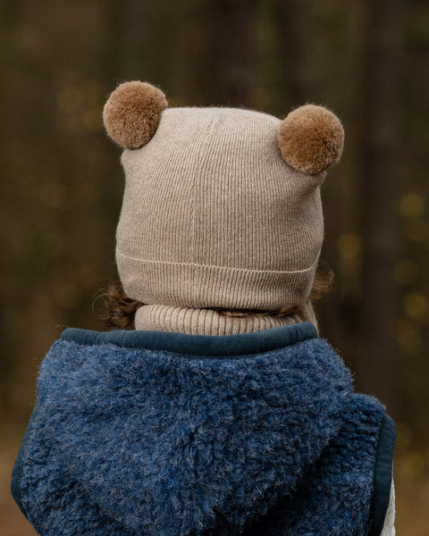 The back of a young child's head and shoulders are shown, with the child wearing a beige knit hat with two brown pom-pom ears. They also have a beige turtleneck and a dark blue fuzzy vest on, with a blurred outdoor background.