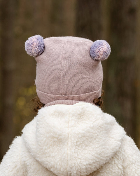 The back of a child's head and shoulders are shown, with the child wearing a light pink knit hat adorned with two pink and purple speckled pom-poms on top. They also wear a lighter pink turtleneck and the hood of a cream-colored sherpa jacket is visible, set against a blurred forest background.