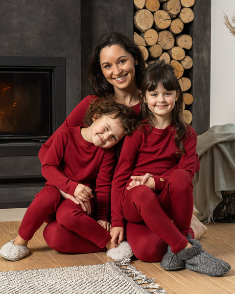 A family of three, a woman and two children, are sitting on a light-colored wooden floor in front of a modern fireplace with a fire burning, and a stack of firewood beside it. All three are wearing matching menique long-sleeved, deep red pajama sets. The woman is in the back, smiling directly at the camera.