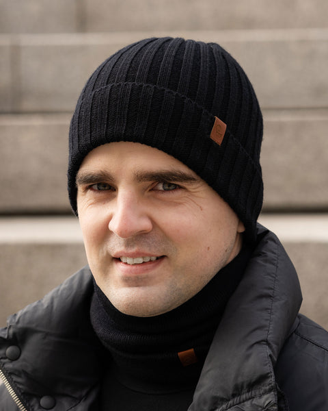 A person with light skin and short dark hair is looking towards the camera with a slight smile. They are wearing a black ribbed knit beanie with a small brown tag, a black turtleneck sweater, and a black puffer jacket. The background appears to be concrete steps in an outdoor setting.