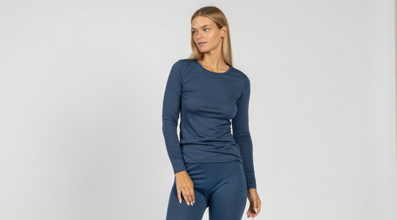 A young blond woman wearing a full set of merino wool base layer clothing of 160gsm fabric in denim color. The set includes a long sleeve shirt and pants. She is looking sideways. 