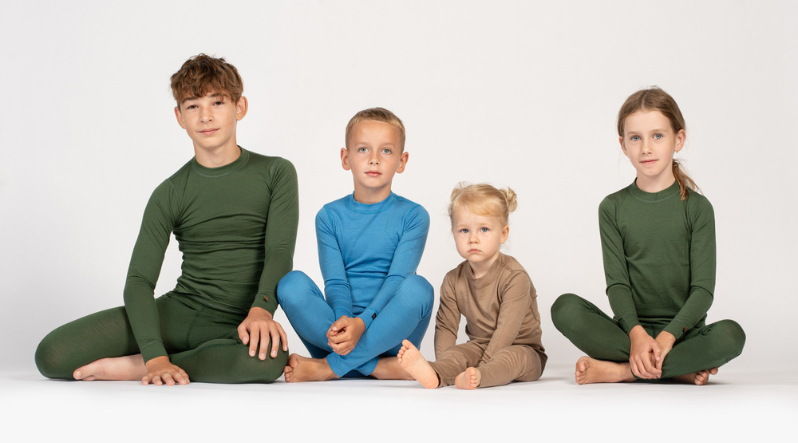 Four kids sitting down, wearing merino wool base layers of 160gsm fabric. The oldest boy wears a dark green set, as well as the oldest girl. The younger boy wears a light blue set, and the baby toddler girl is wearing a creamy beige one.