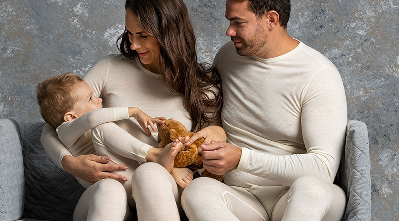 In the photo you can see a family, where mom and dad is looking at their child. They are all wearing natural color two-piece pajamas/loungewear sets made from warm and organic 100% Merino wool.
