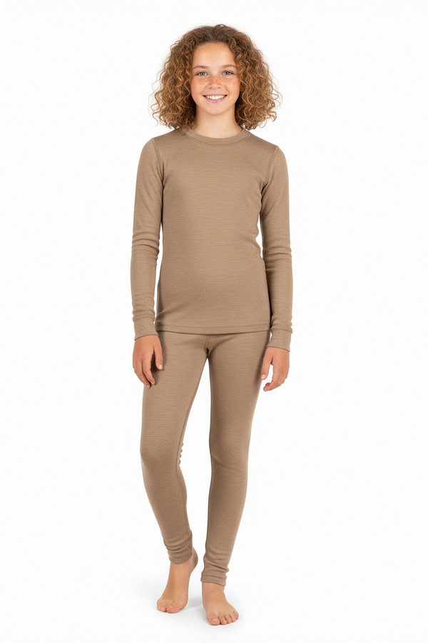 A girl wearing a beige long-sleeve Merino wool base layer top and matching leggings, standing barefoot and smiling against a white studio background.