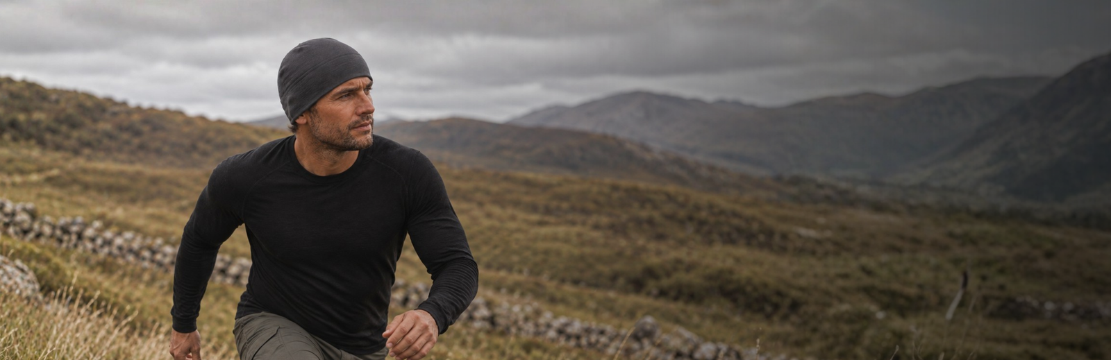 A man wearing a black long-sleeved thermal top and a dark grey beanie hikes up a rugged, grassy hillside. He looks intently off to the side, set against a vast backdrop of rolling mountains and a dark, moody overcast sky.