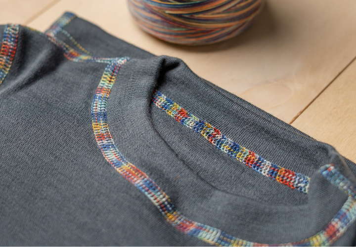 Close-up of a dark gray merino wool garment from menique, showcasing colorful flatlock stitching along the collar and seams, with a multicolored thread spool in the background.