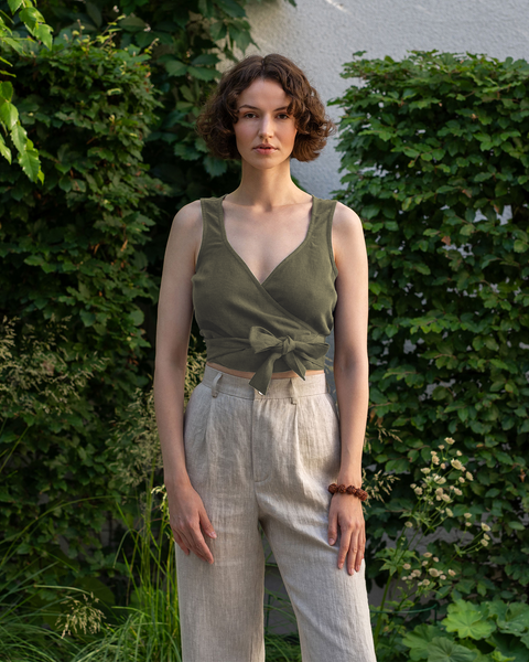 A woman wearing 100% linen sleeveless wrap crop top in the color stone green. She is looking straight and the photo&#39;s background is green and full of vegetation. 