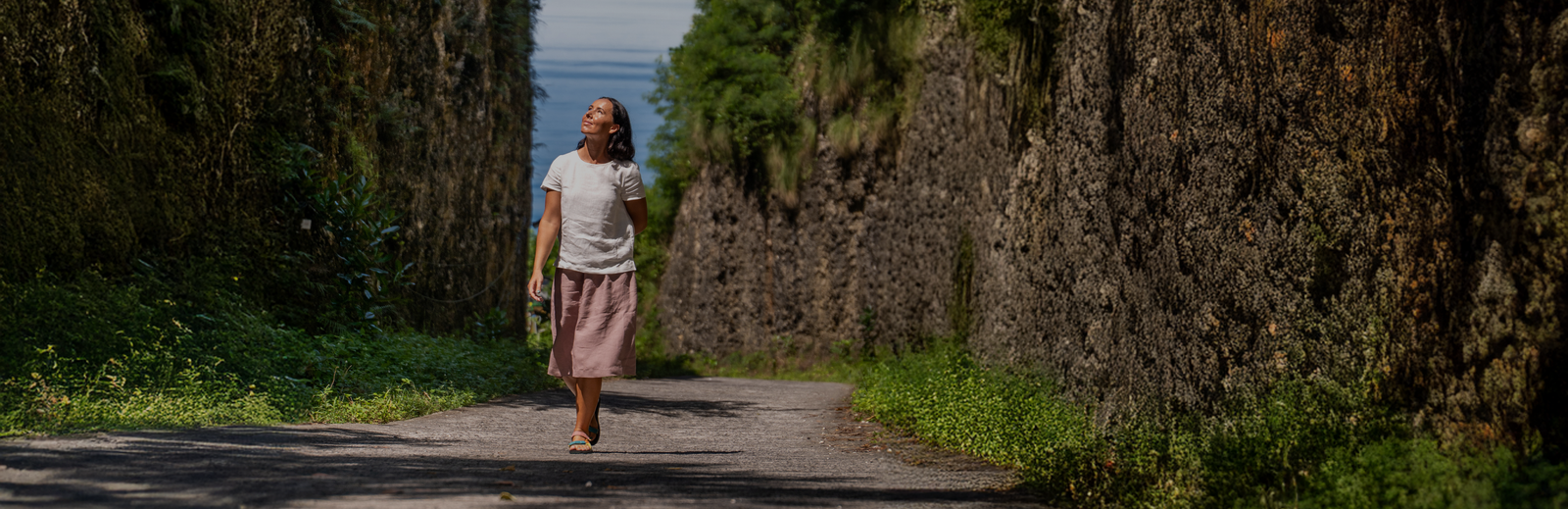 A woman walks down a paved pathway nestled between two high, moss-covered stone walls, looking up towards the sky. She is dressed casually in a white short-sleeve top and a loose, dusty pink skirt.